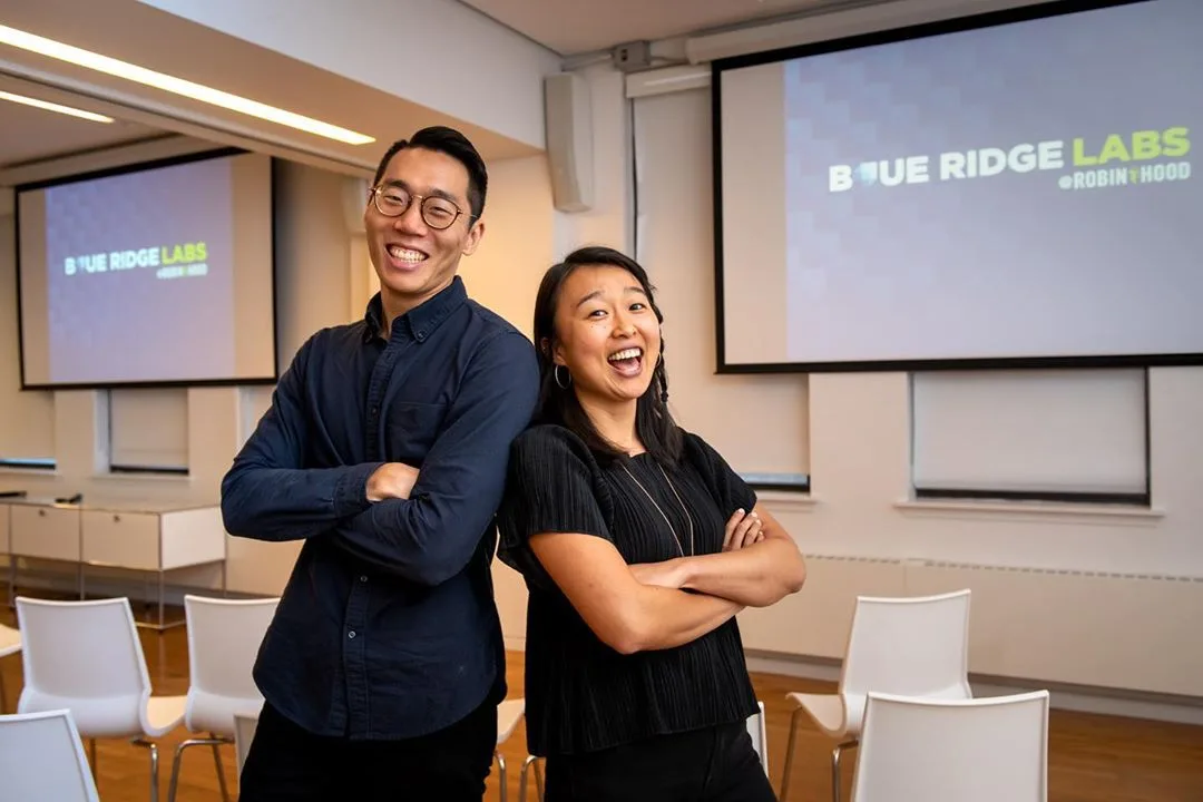 Two people posing back-to-back in front of two large display screens featuring Blue Ridge Labs and Robin Hood's logos.