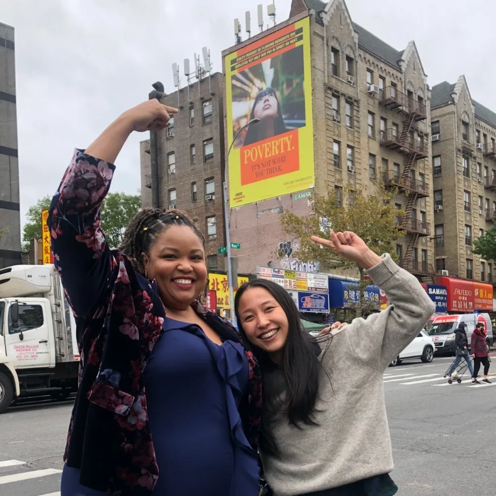 Two women posing in an outdoor setting and pointing towards a large advertisement on a building exterior